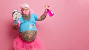 Cleaning-Tips-Horizontal shot of bearded man has big tattooed belly holds spray detergent and plunger wrapped with toilet paper wears fairy costume isolated over pink background with copy space. Housekeeping