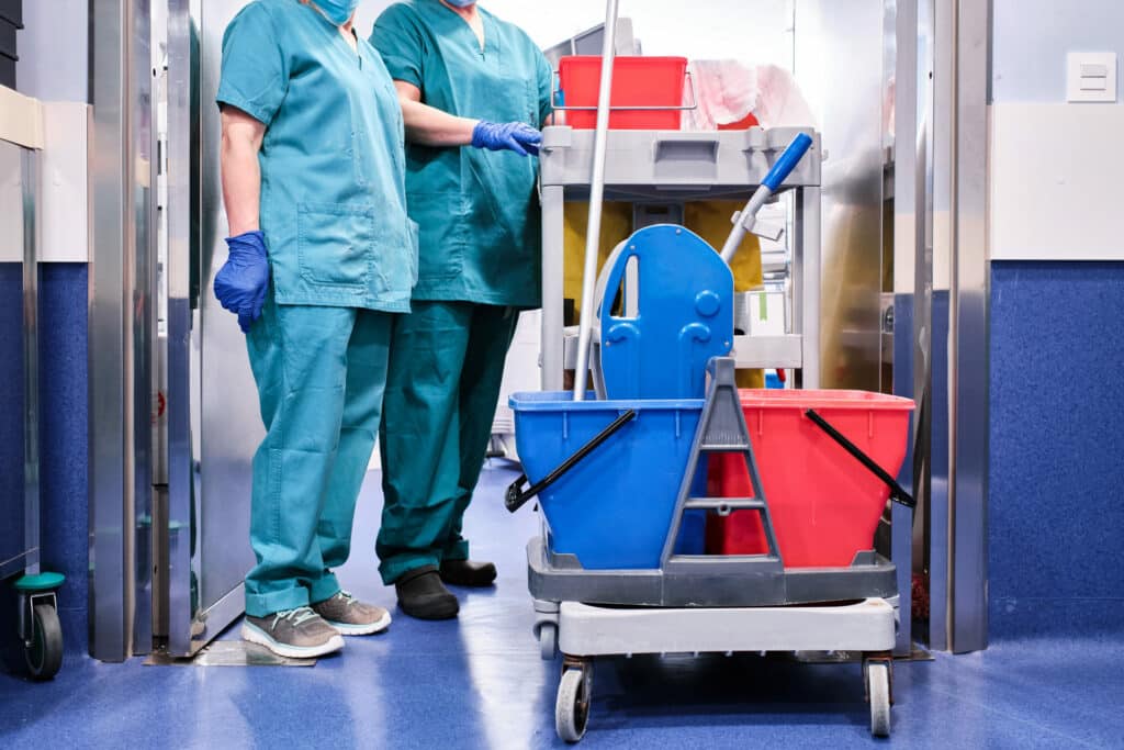 How to Hire a Professional Cleaning Company. 2 cleaners standing next to cleaning supplies in hospital gowns.
